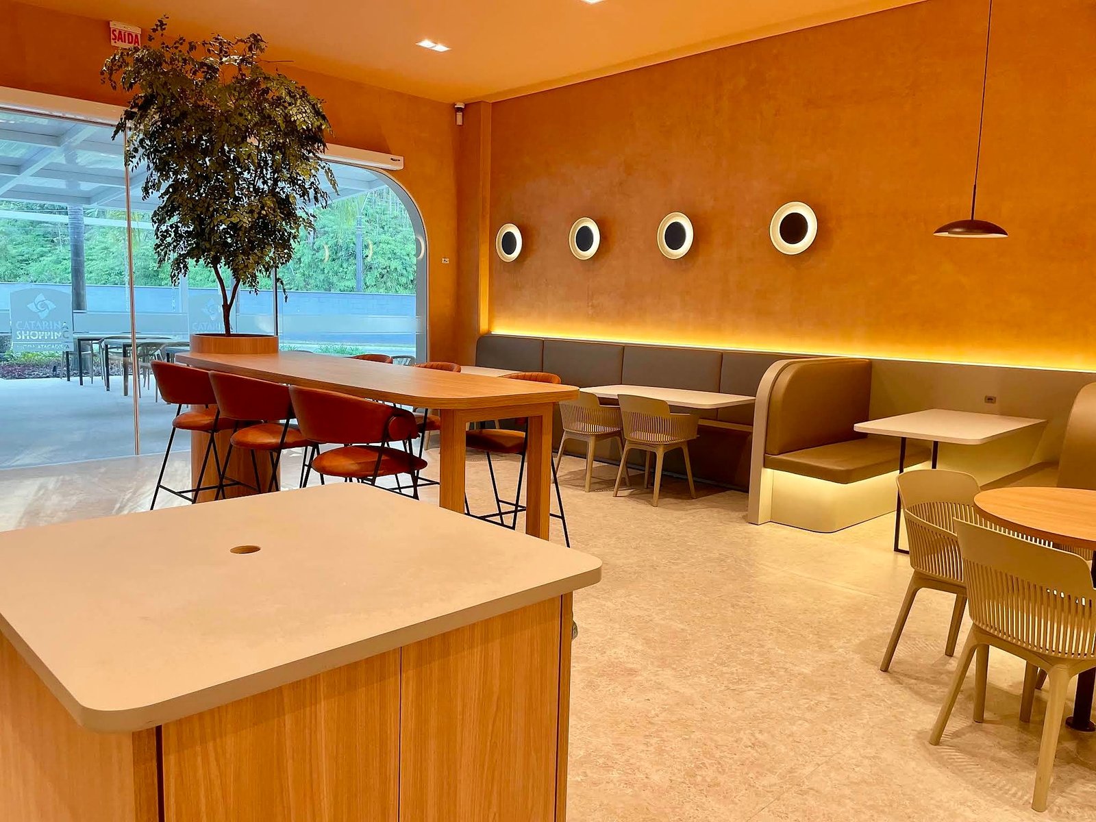 Salão da Brisa Café, com paredes terracota, luz embutida, banco estofado e janela em arco para o garden.